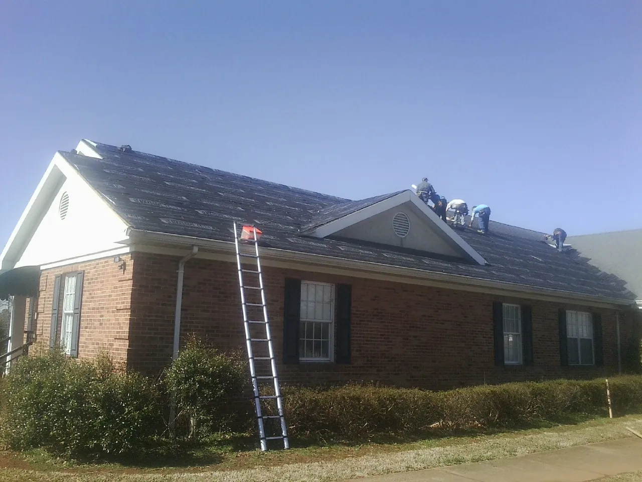Crew tearing shingles off a brick ranch roof with a ladder set — Emergency Roof Repair work by our crew in Lake Placid
