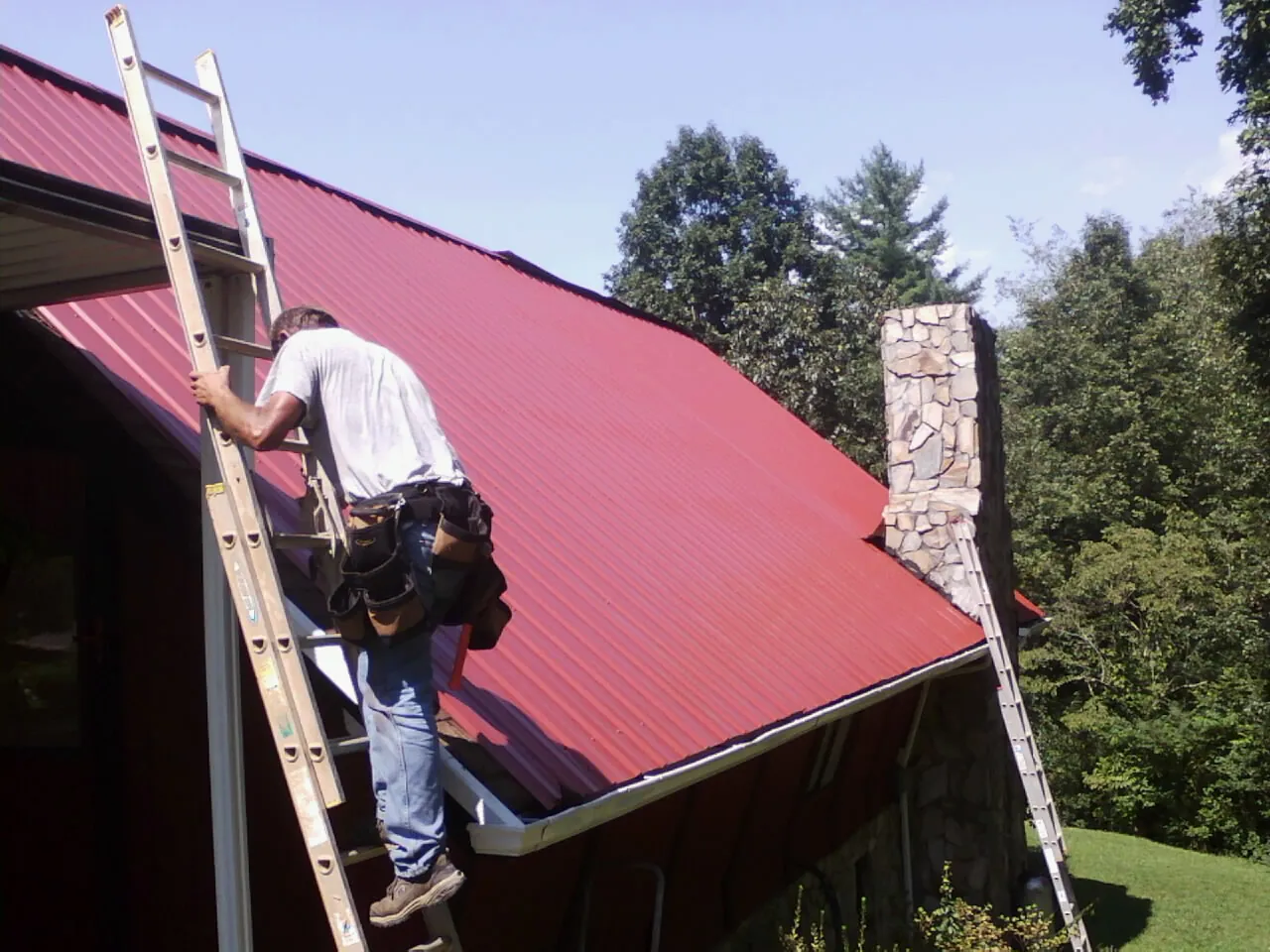 Roofer on a ladder beside a red metal roof and stone chimney — Soffit & Fascia Repair by Roofing Brothers in Lake Placid